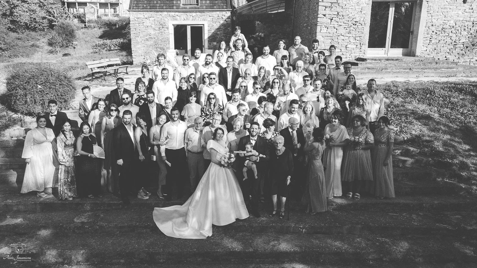 Groupe de personnes lors d'un mariage en extérieur, avec la mariée en robe blanche au centre, entourée de la famille et des amis, en tenue formelle, devant un bâtiment en pierre.