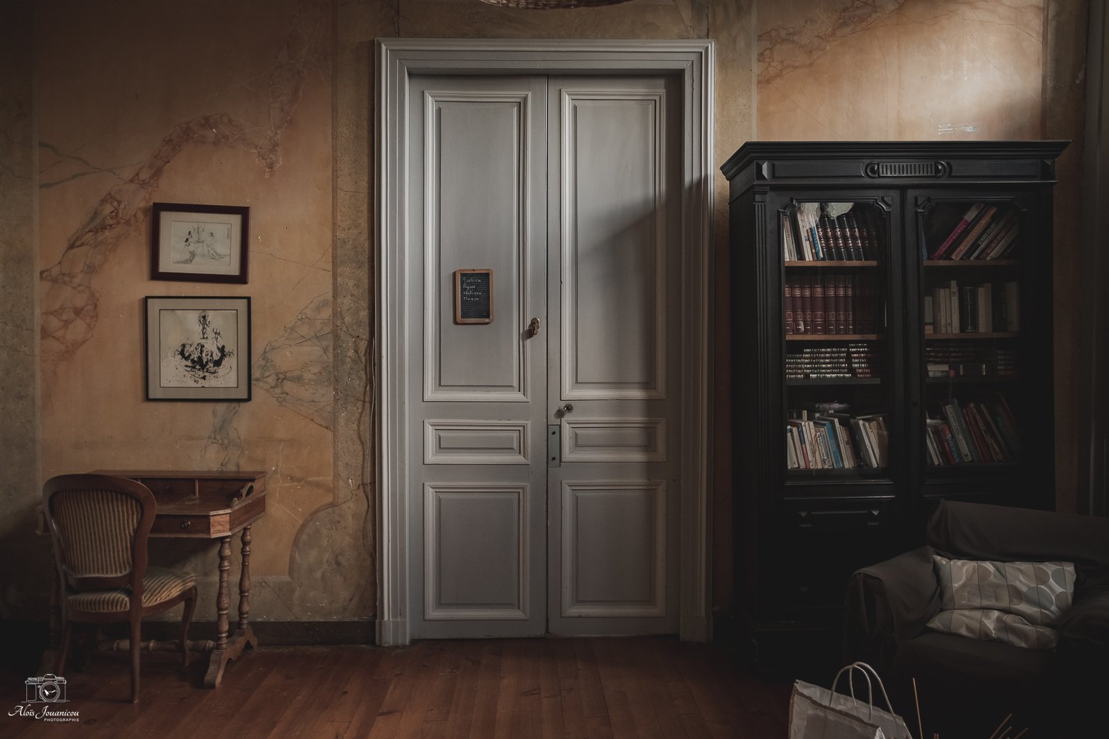 Intérieur d'une pièce avec une porte en bois gris clair, un meuble en bois, une étagère noire remplie de livres, deux cadres avec dessins sur le mur, un fauteuil en bois avec un coussin et un sac à côté.