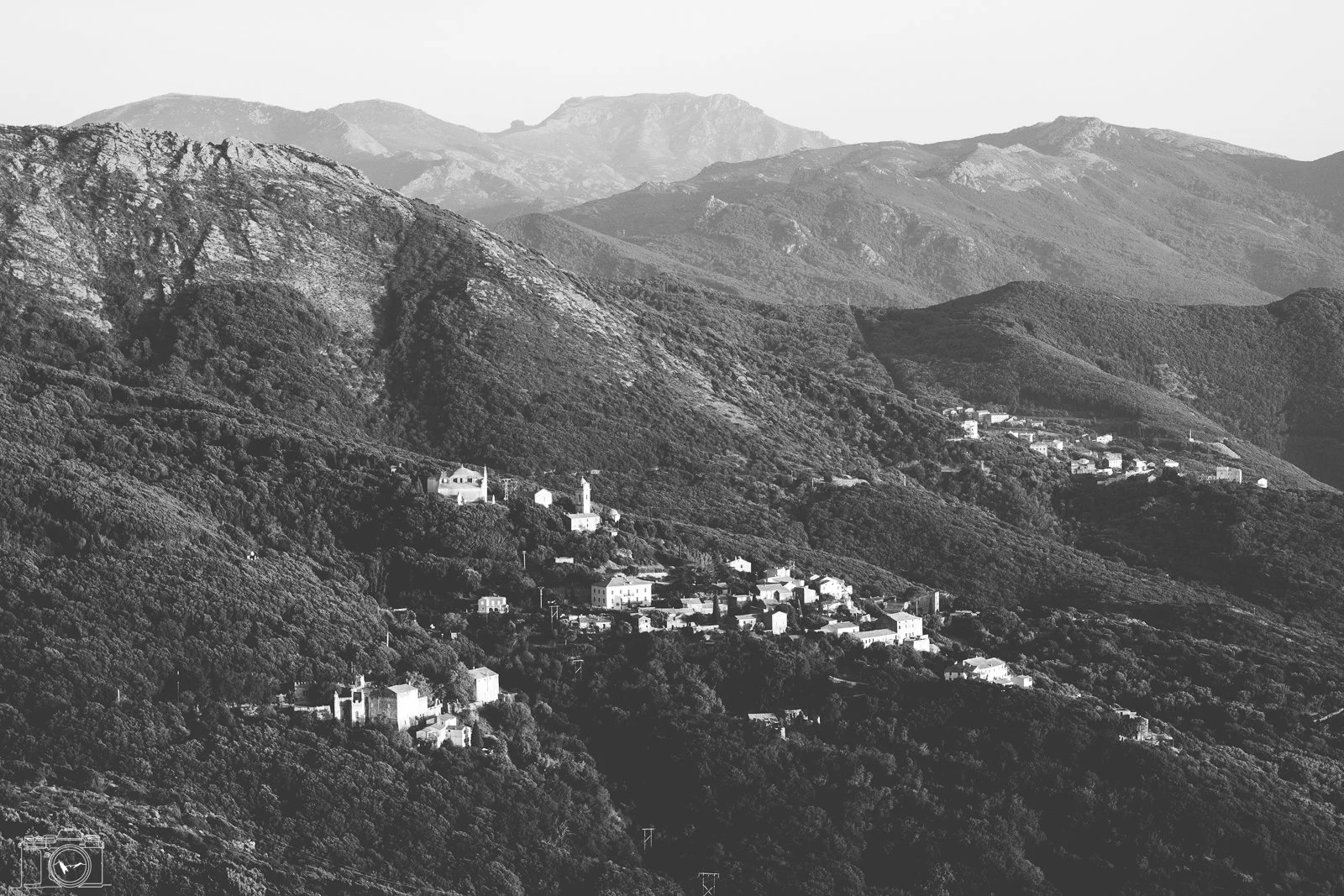 Paysage de montagnes avec un village accroché sur la pente, en noir et blanc.