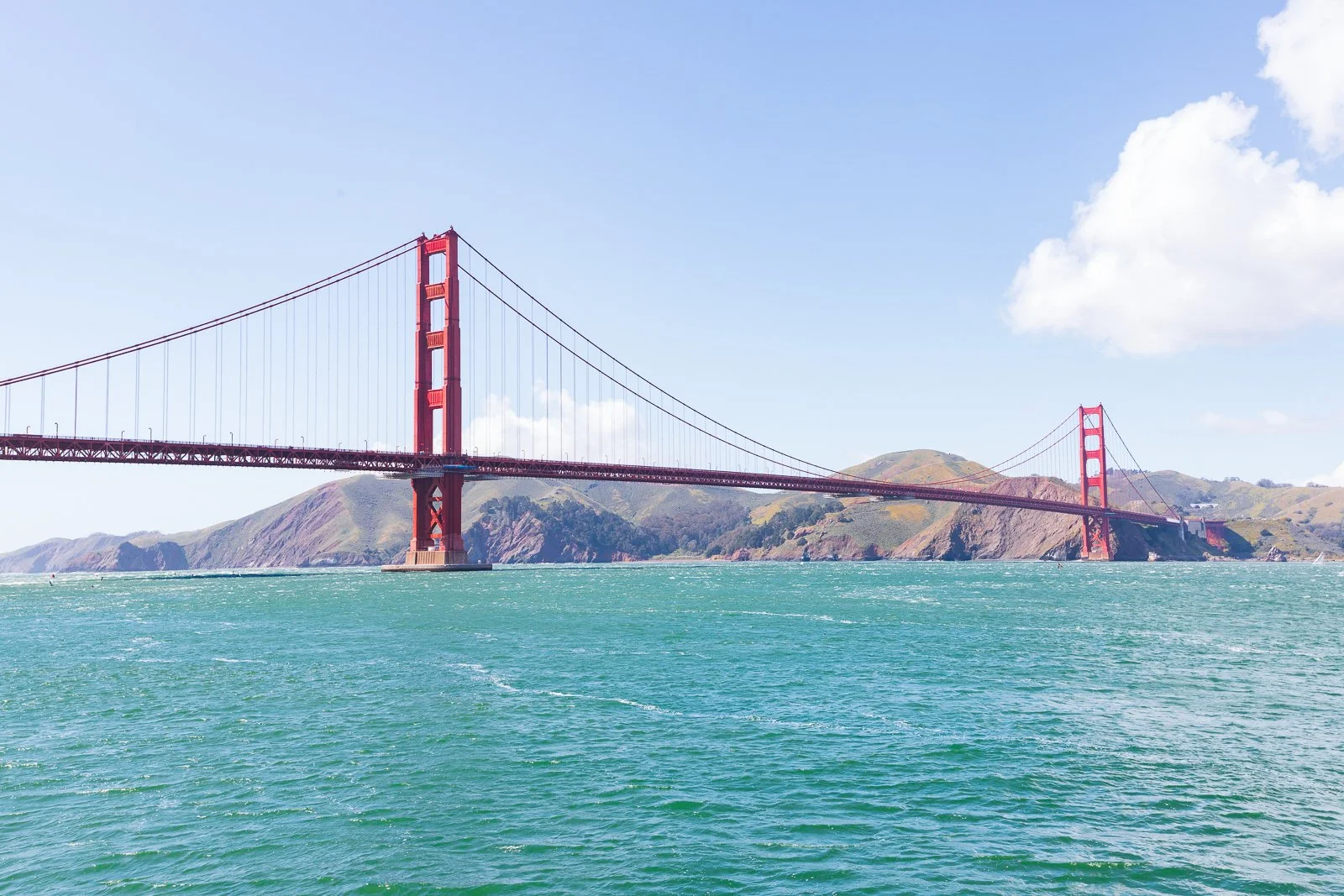 Le pont du Golden Gate avec ses grandes tours rouges, en Californie, avec une mer turquoise et des collines en arrière-plan, sous un ciel bleu avec quelques nuages.