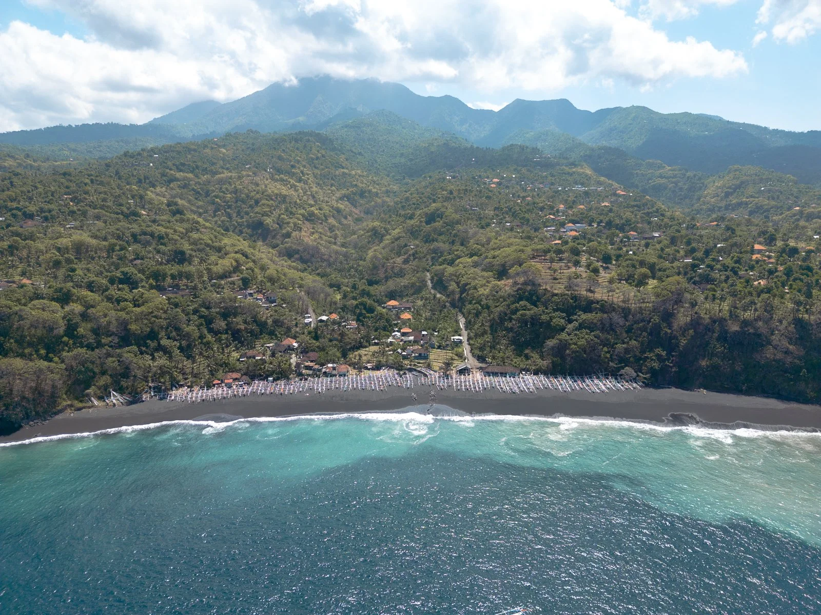 Côte rocheuse avec plage de sable noir bordée de bateaux, forêt verdoyante, montagnes en arrière-plan, ciel nuageux.