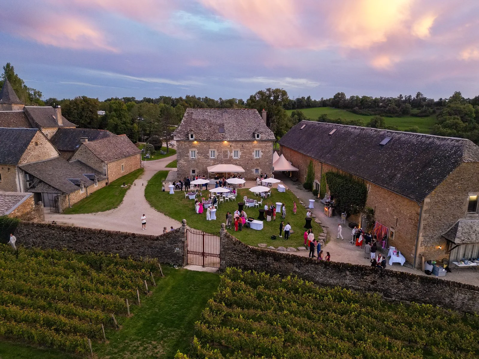Célébration ou réception en plein air dans une cour de ferme médiévale avec des invités, des tables avec parapluies, entourée de bâtiments en pierre, au crépuscule.