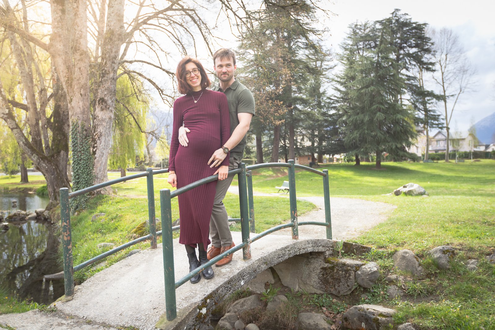 Un couple pose sur un petit pont en pierre dans un parc, la femme est enceinte, la scène se déroule au printemps avec des arbres en fleurs et un ciel partiellement nuageux.