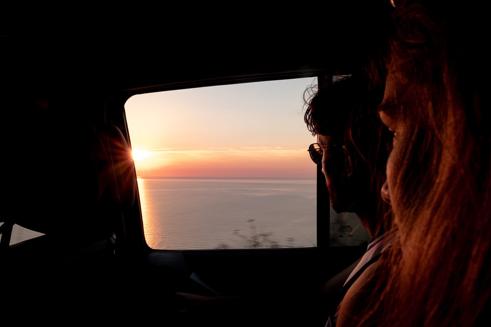 Deux personnes assises à l'intérieur d'un véhicule regardant un coucher de soleil sur l'océan, avec la lumière du soleil se couchant à l'horizon.
