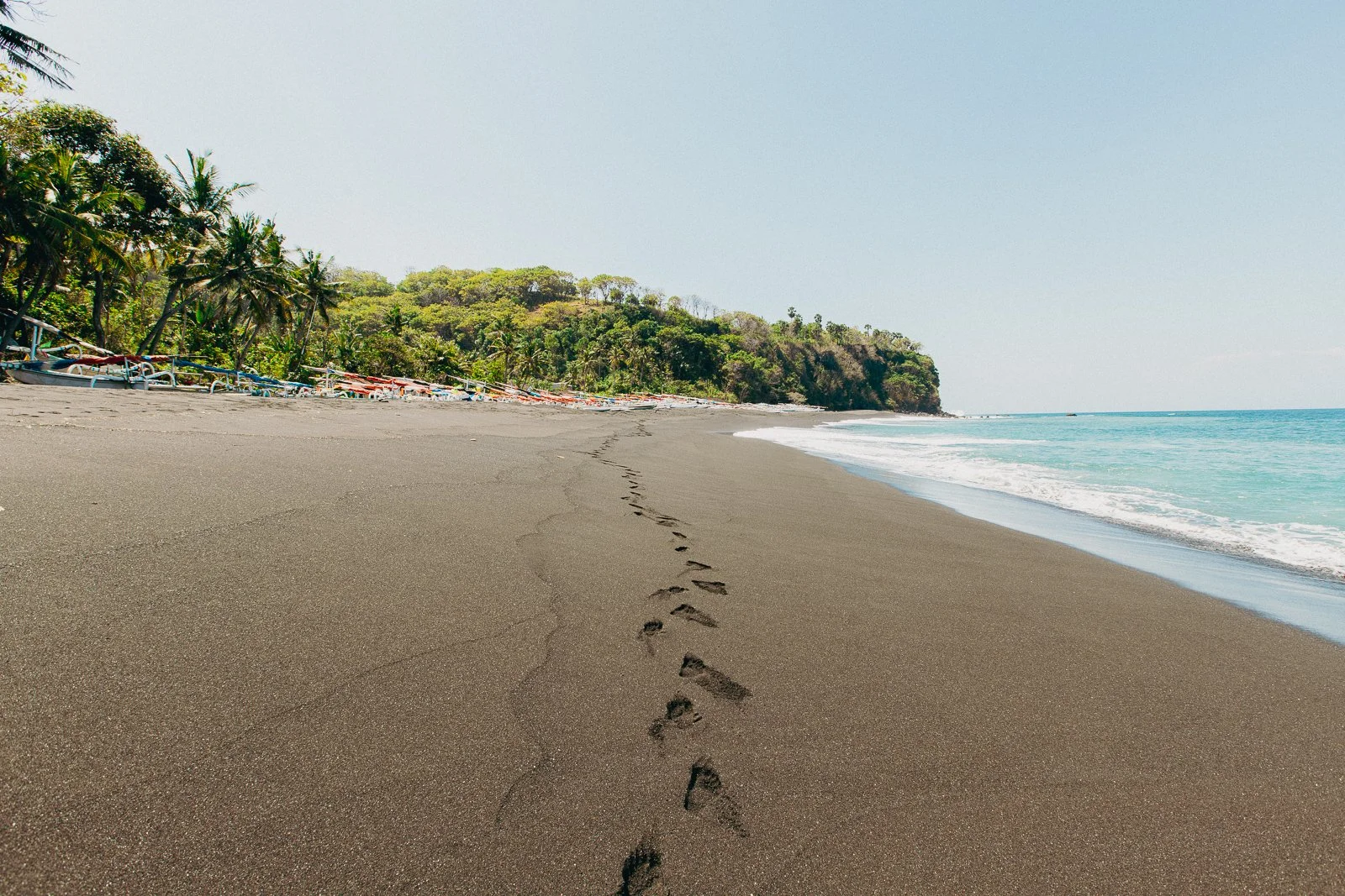 Une plage de sable noir avec des empreintes de pas, des palmiers et un bateau à côté d'une forêt tropicale, face à la mer calme 