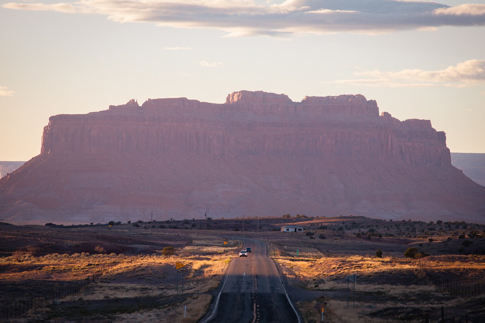Une route droite traversant un paysage désertique avec un immense plateau rocheux en arrière-plan, sous un ciel partiellement nuageux, probablement dans une région désertique ou semi-aride.