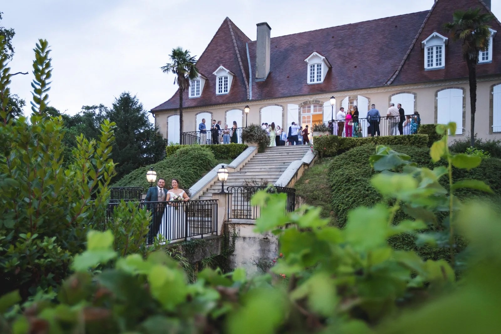 Gens en costume de mariage devant une maison de style traditionnel avec escaliers et jardin bien entretenu.