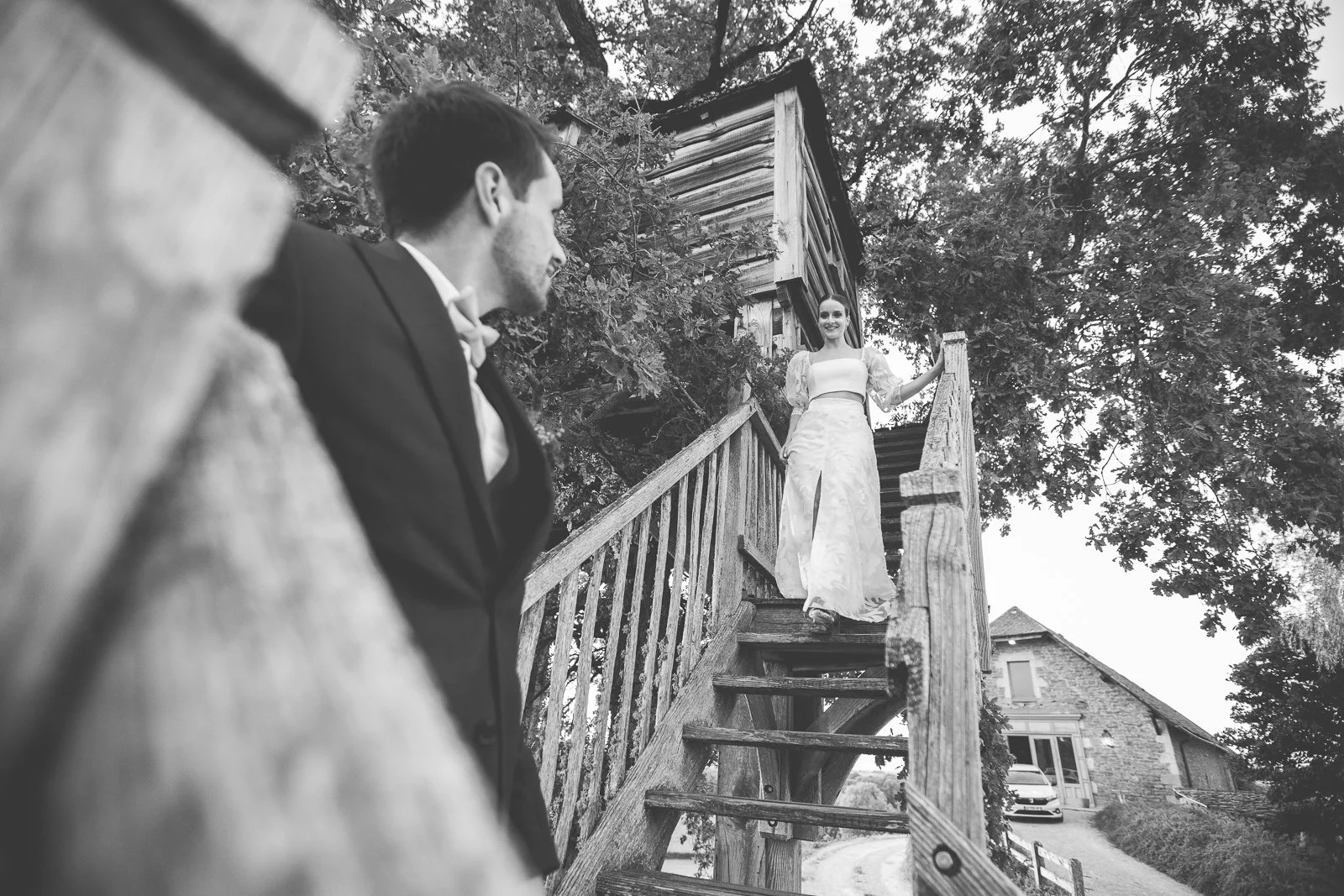 Un homme en costume regardant une femme en robe de mariée qui se tient sur un escalier en bois en extérieur, avec des arbres et une maison en arrière-plan.