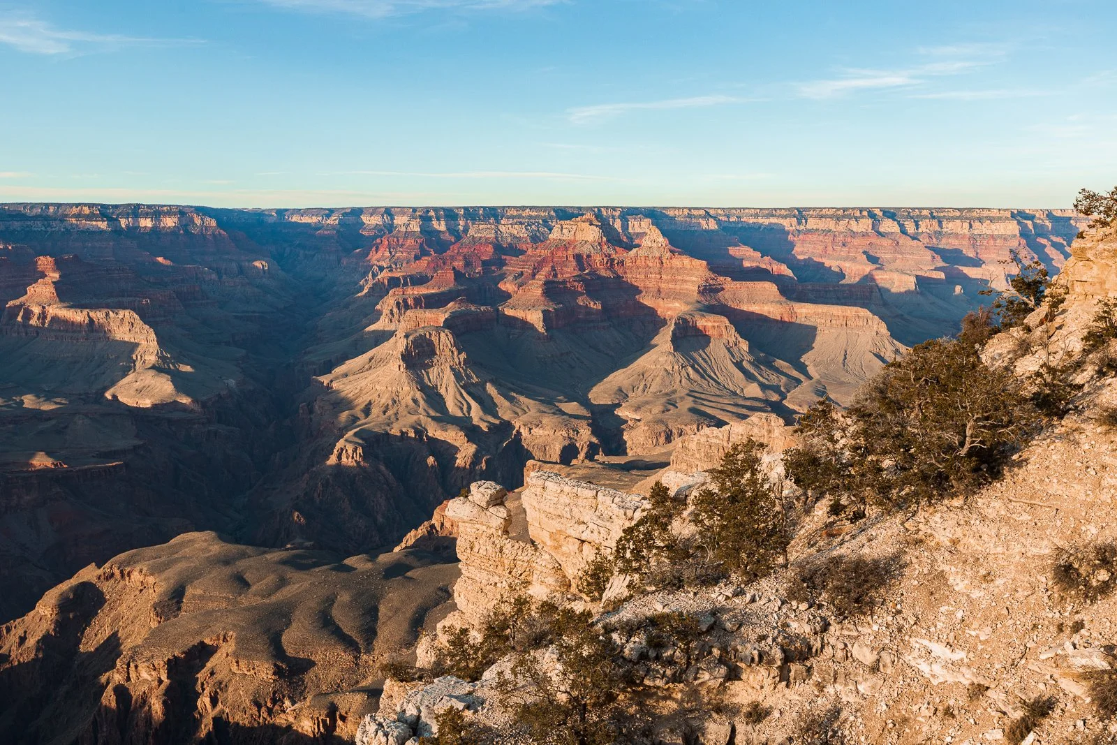 Vue panoramique du Grand Canyon avec des formations rocheuses colorées et un ciel clair, au coucher du soleil.