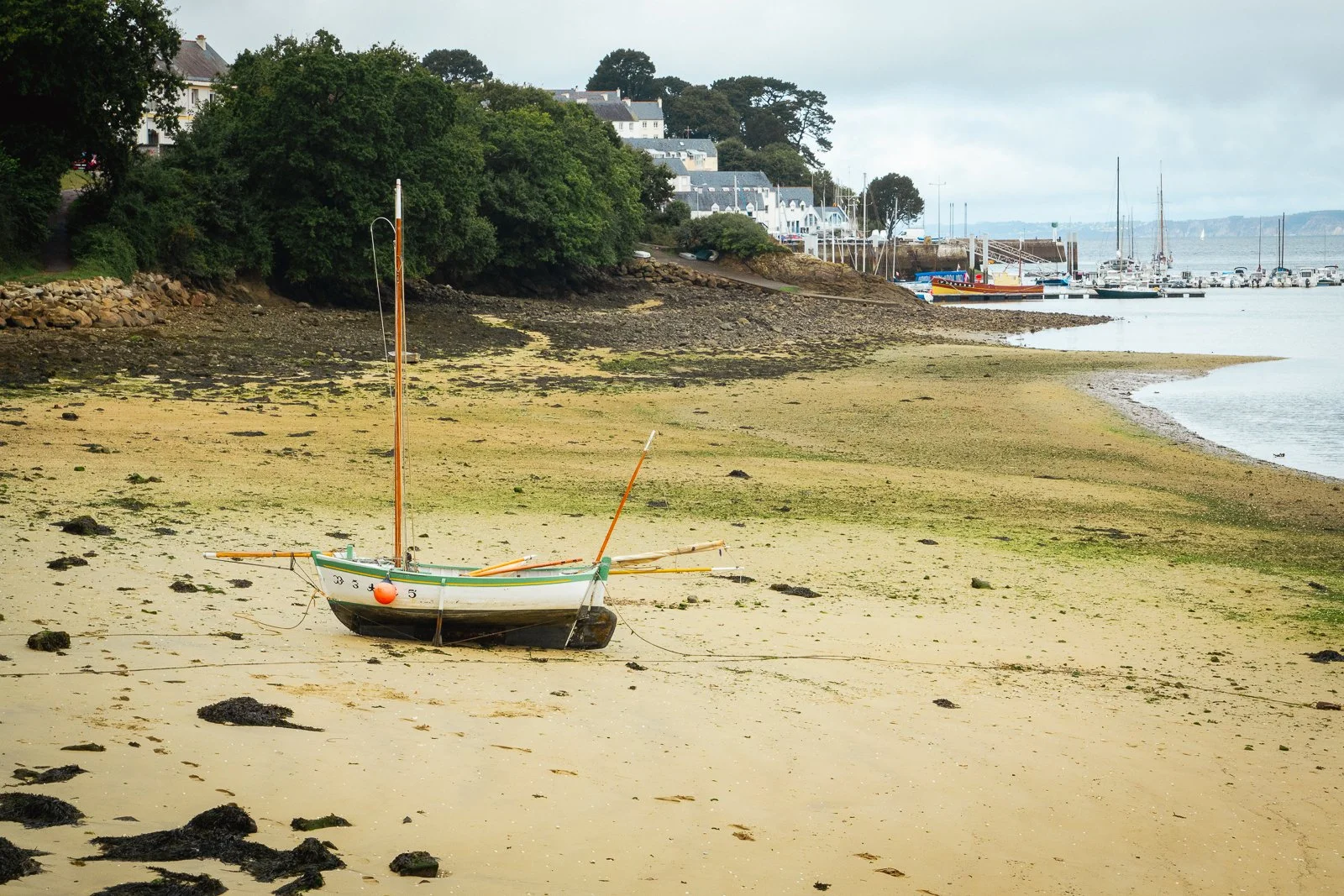 Un bateau de pêche échoué sur une plage de sable, avec des maisons et un port de yachts en arrière-plan, sous un ciel nuageux.