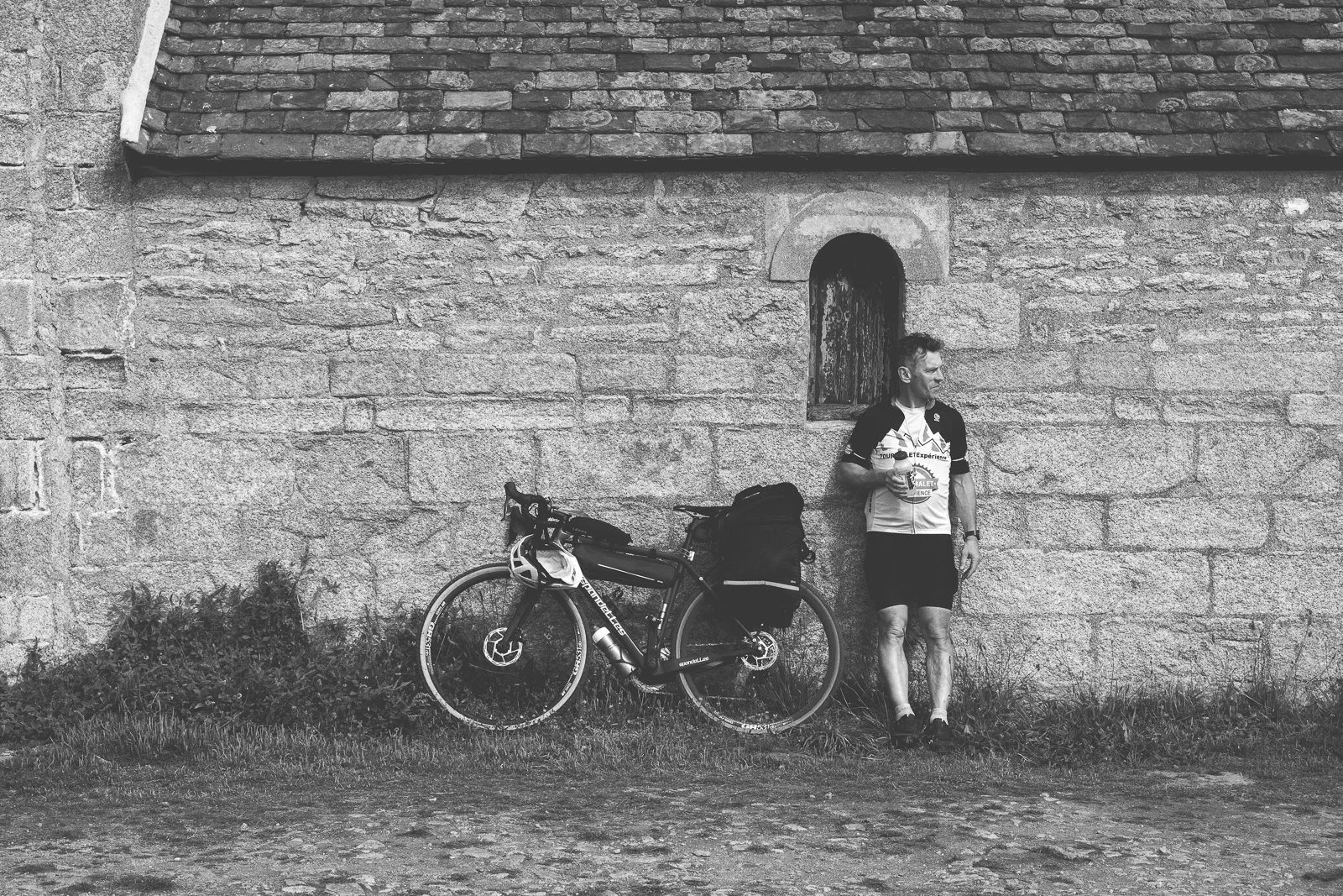 Homme cycliste en tenue de sport, avec vélo chargé, buvant dans une tasse, qui se repose contre un mur en pierre ancienne.