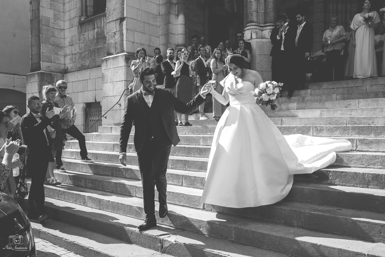 Un couple de mariés descend les escaliers d'une église, entouré d'invités, en noir et blanc.