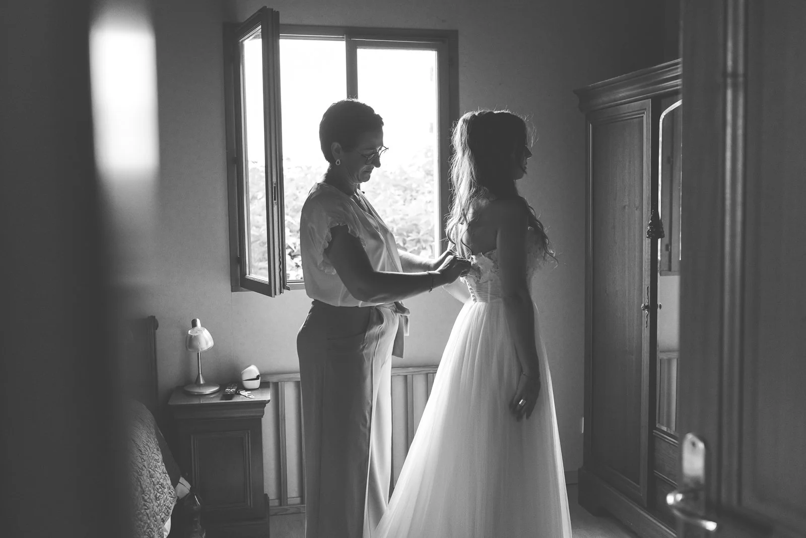 Une femme plus âgée ajustant la robe de mariée d'une jeune femme dans une chambre, avec une fenêtre ouverte laissant entrer la lumière naturelle.