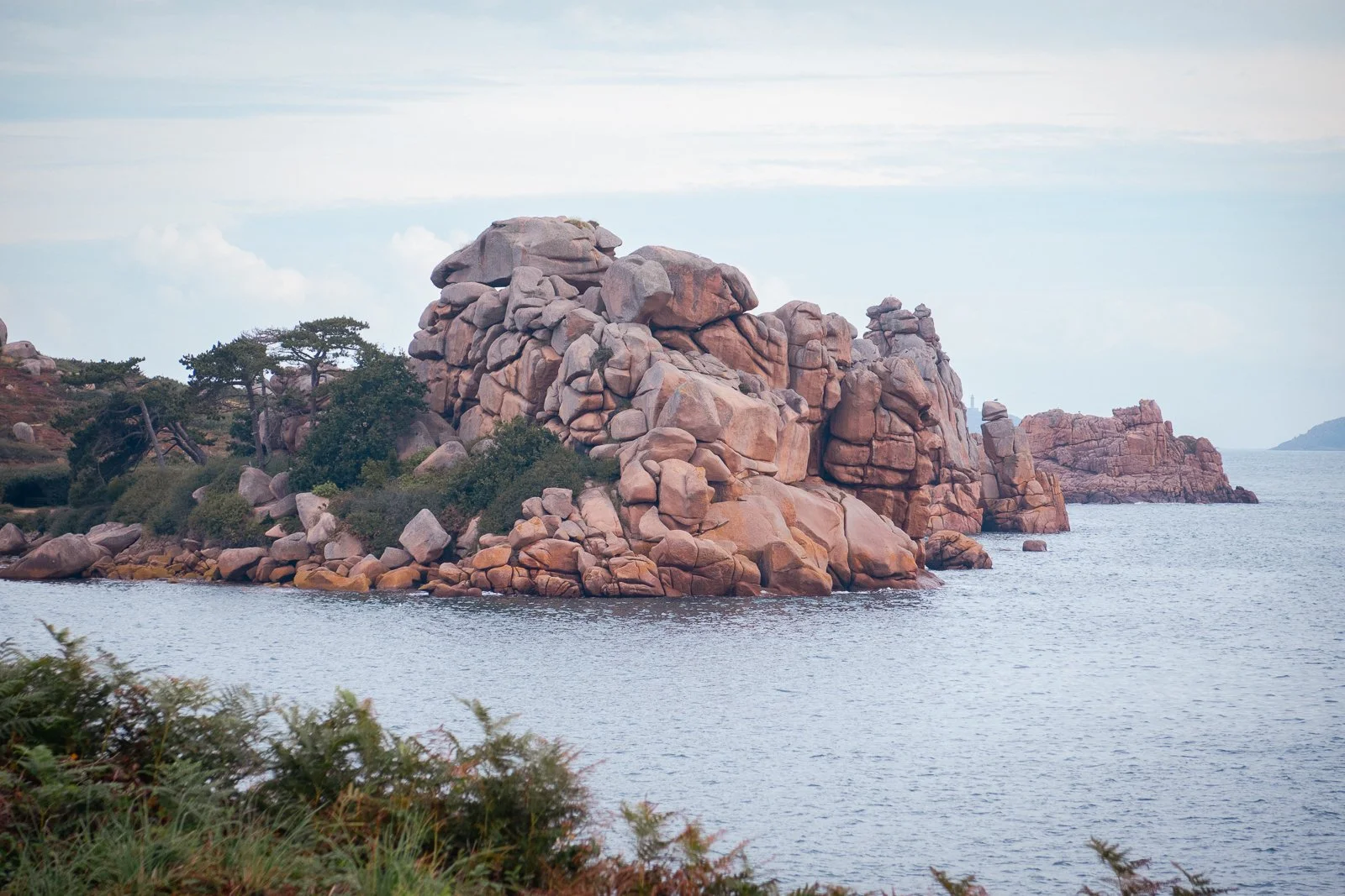 Un paysage côtier avec des rochers géants et quelques arbres, vue sur la mer et des îles lointaines.