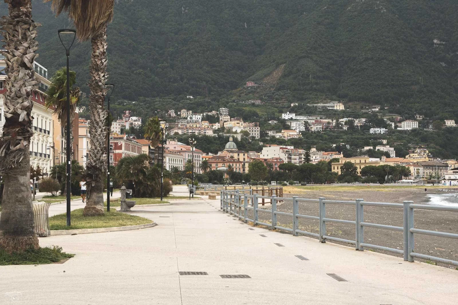 Une promenade en front de mer avec des palmiers, des lampadaires, des bâtiments colorés et des collines verdoyantes en arrière-plan.