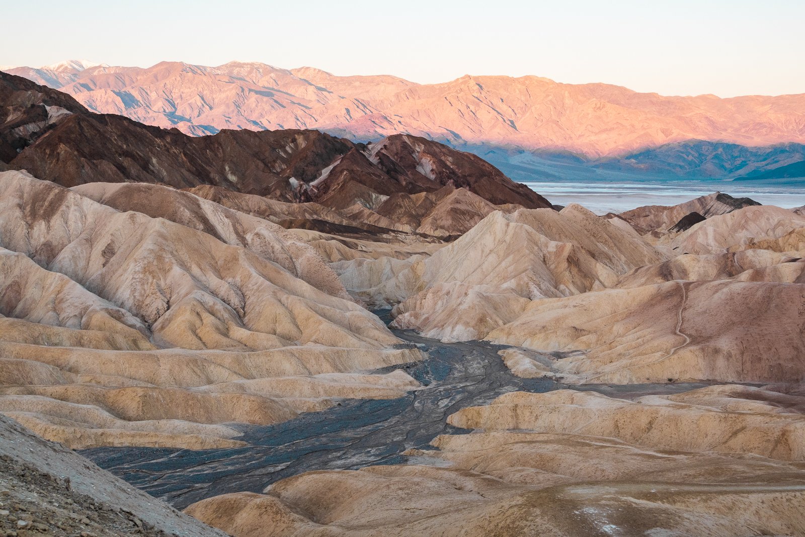 Paysage désertique avec montagnes colorées en arrière-plan, un lac ou une étendue d'eau, et un lit de rivière asséché avec des formations rocheuses en teintes beige, marron et grise.