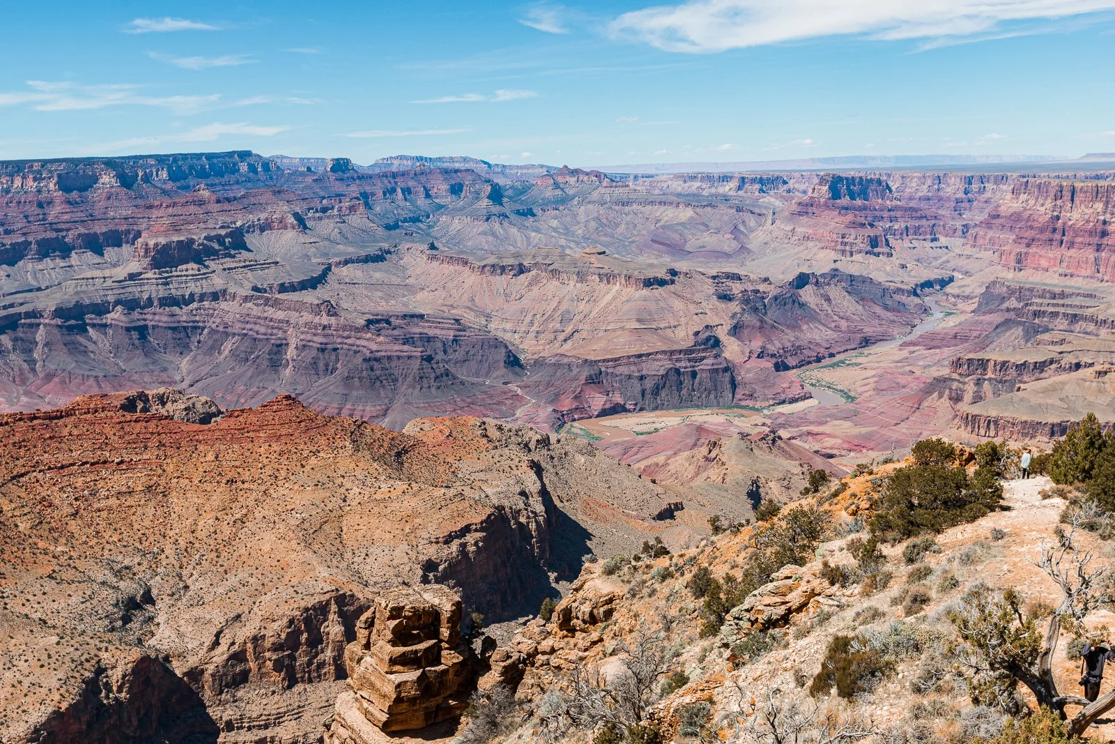 Vue panoramique du Grand Canyon avec ses couches rocheuses colorées, le fleuve Colorado en contrebas, et un ciel bleu clair avec quelques nuages.