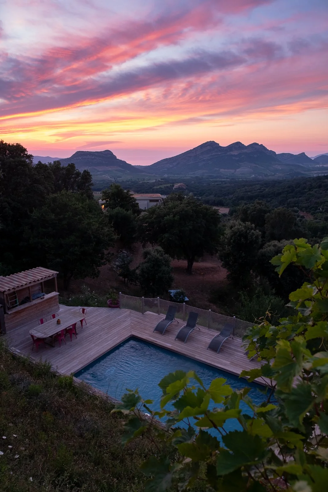 Une vue de coucher de soleil sur des montagnes, avec un ciel coloré en nuances de rose, orange et violet, au loin. En premier plan, une piscine avec des chaises longues autour et un espace de détente, entourée de végétation et de collines verdoyantes