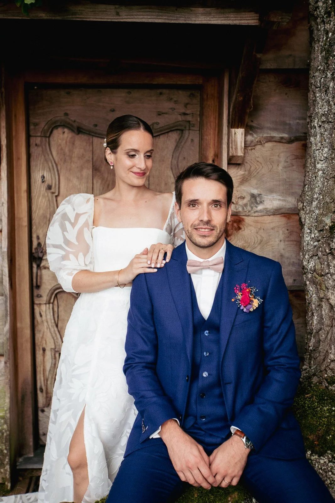 Un couple en tenue de mariage pose devant un décor en bois. La femme en robe blanche et le homme en costume bleu. Elle a les mains sur son épaule.