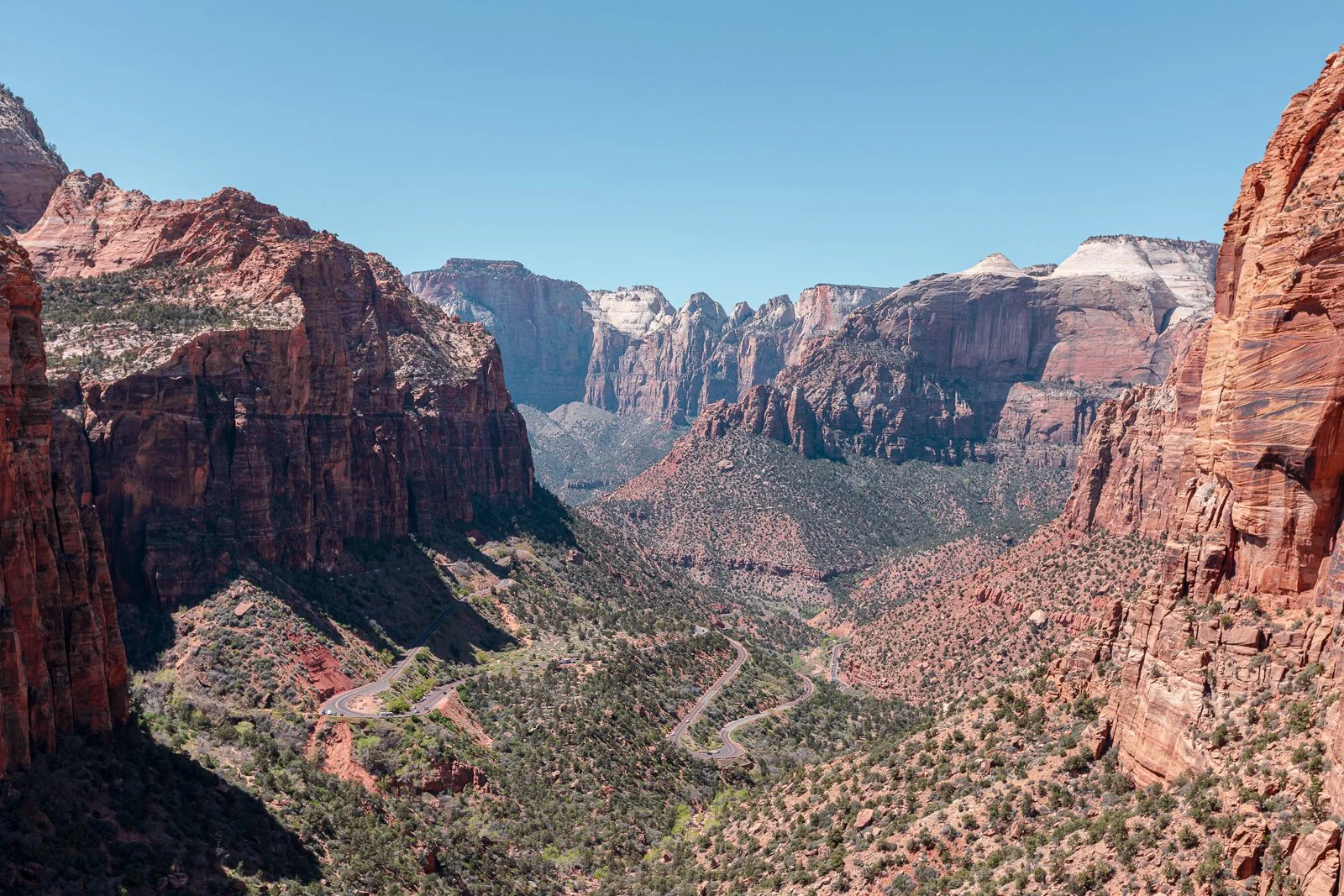 Canyon avec des rochers rougeâtres, un ciel bleu clair et des montagnes enneigées en arrière-plan, avec une route sinueuse dans la vallée.