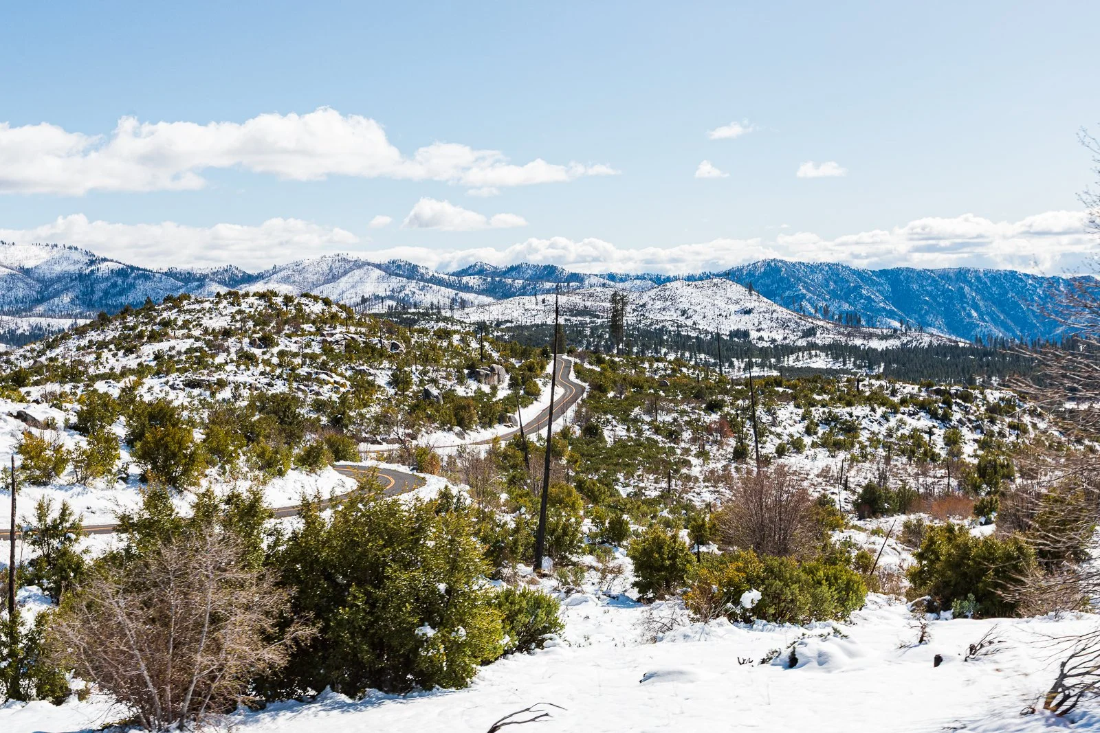 Paysage de montagnes enneigées avec une route serpentant à travers une végétation hivernale dense, ciel partiellement nuageux.