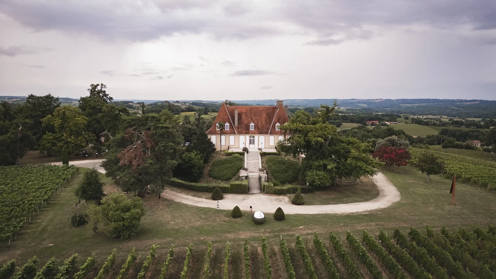 Château pittoresque au centre d'un vignoble entouré de vergers, sous un ciel nuageux.