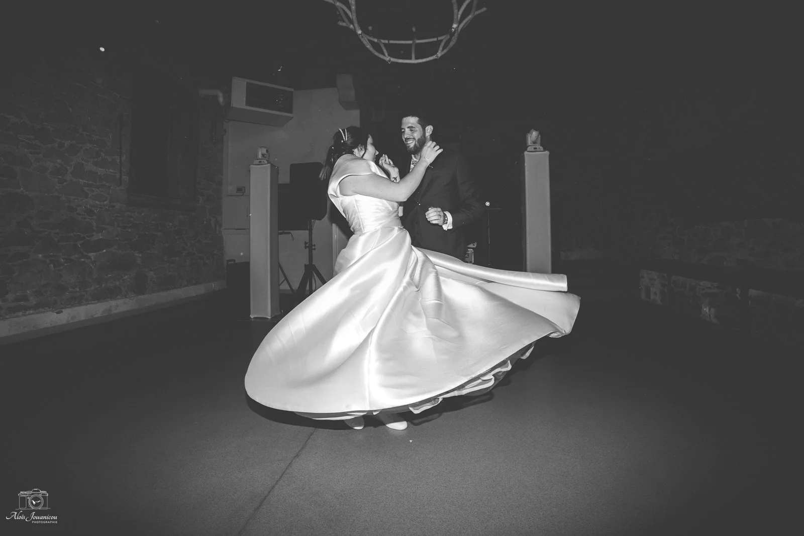 Un couple de mariés danse lors de leur mariage, la mariée porte une robe de mariée fluide et le marié une tenue noire, dans une salle avec murs en pierre et un éclairage tamisé.