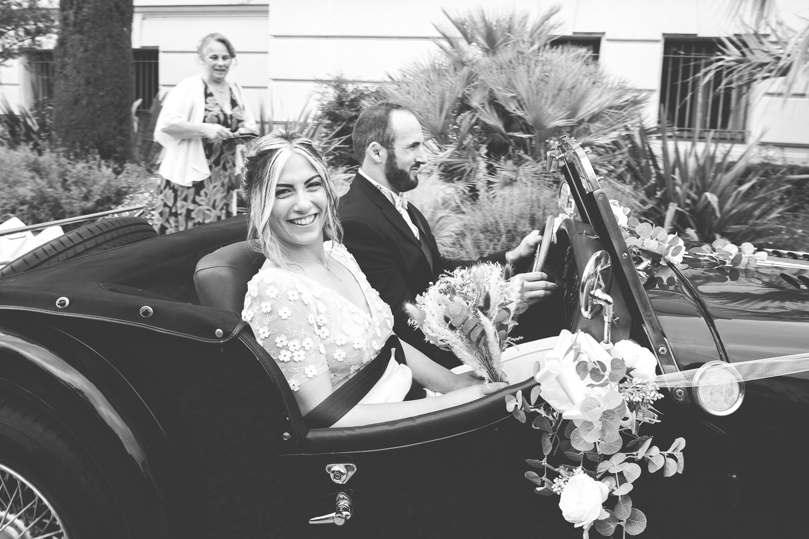 Un couple de mariés dans une voiture vintage décorée de fleurs, avec une femme qui sourit et un homme qui conduit, en arrière-plan une femme qui regarde, dans un cadre urbain.