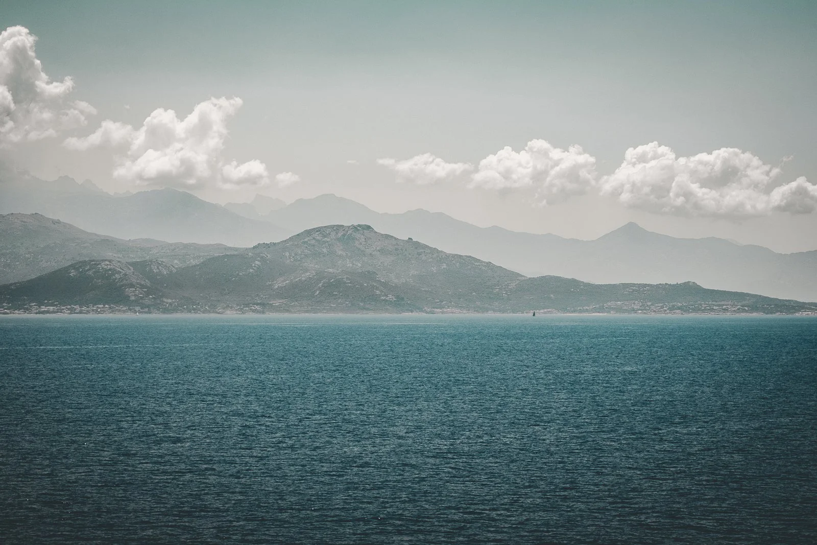 Paysage de montagnes au loin avec un lac ou une mer calmes au premier plan, ciel partiellement nuageux.