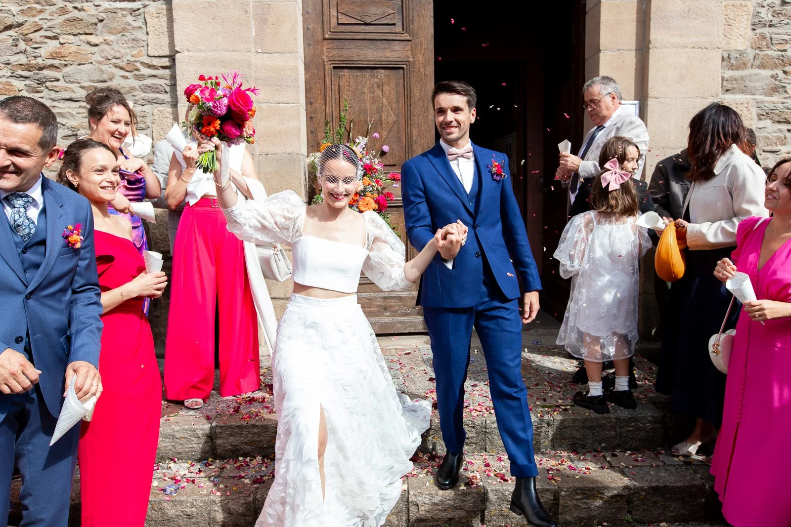 Un couple de mariés souriants sortent de l'église, entourés de leurs invités qui leur lancent des pétales.