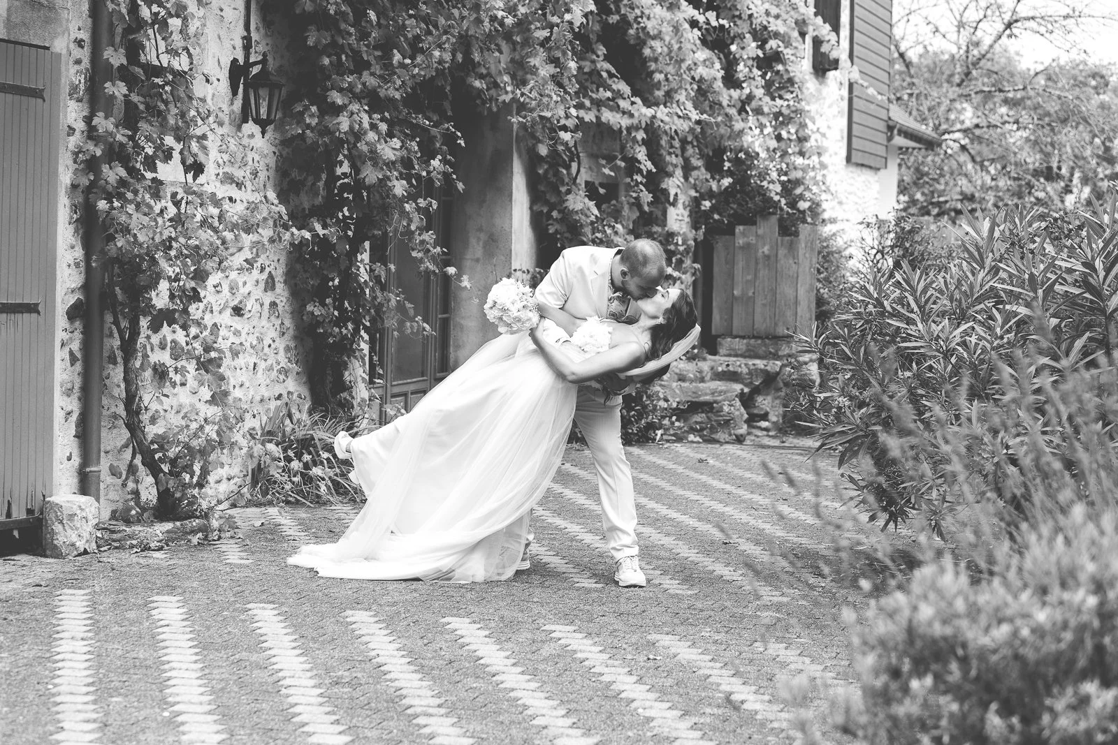 Un couple de mariés en robes blanches, l'homme en costume clair, partage un moment romantique en extérieur, le marié tenant la mariée qui penche légèrement en arrière, dans un cadre bucolique avec des plantes et une vieille bâtisse en pierre.