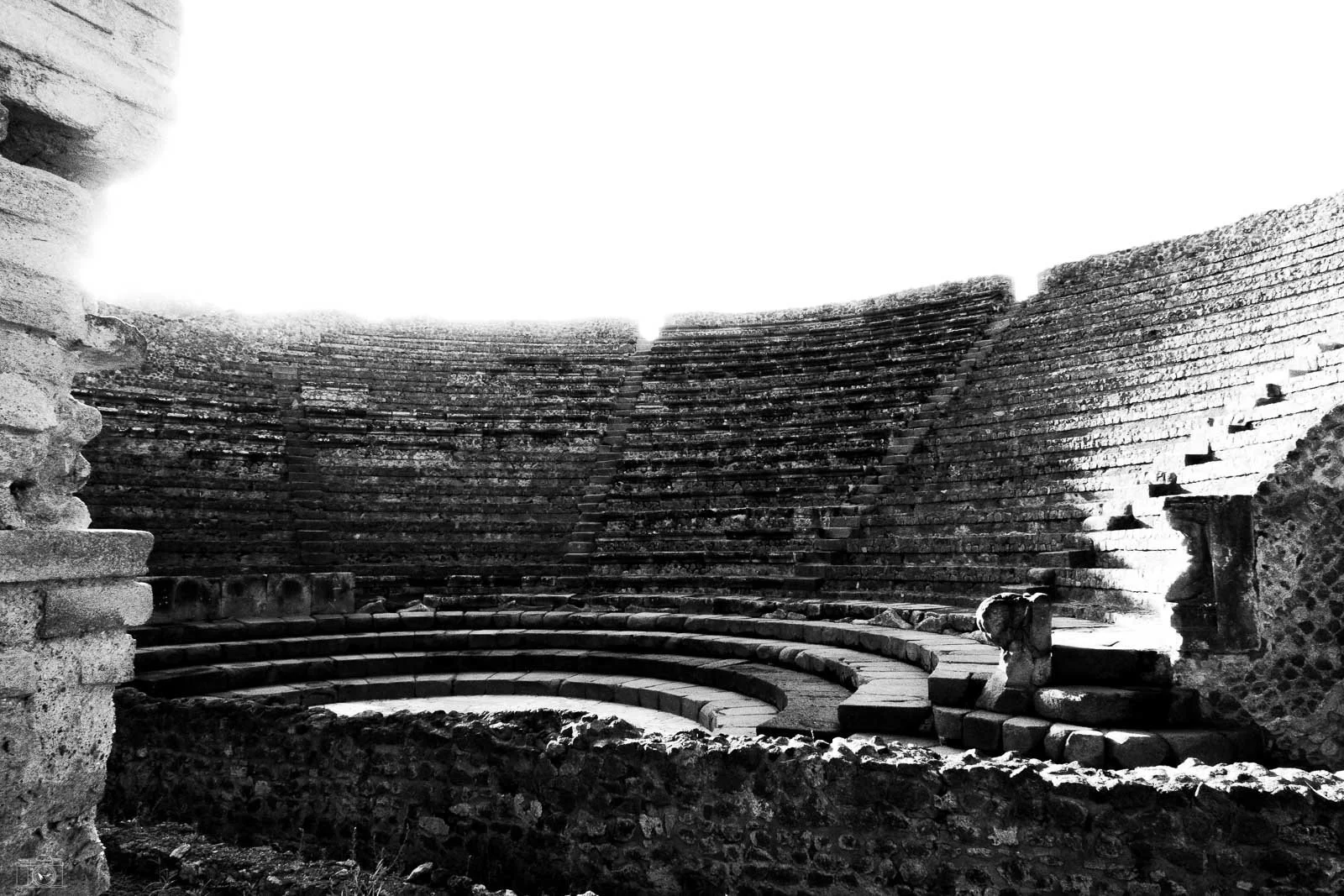 Ancienne arène romaine en pierre, vue intérieure avec gradins en demi-cercle et escalier central, en noir et blanc.