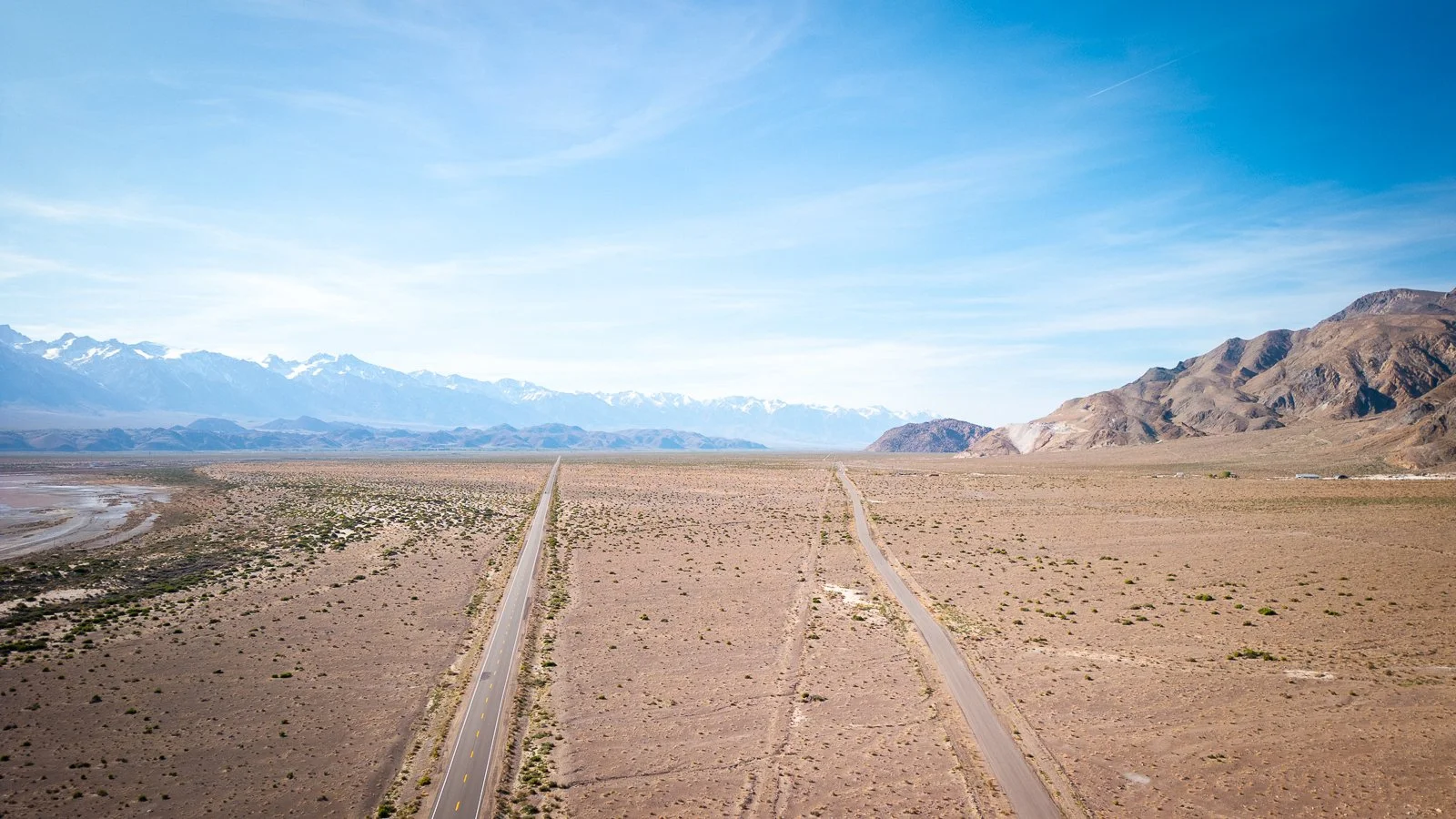 Une image d'un désert avec deux routes pavées traversant la paysage plat, avec des montagnes enneigées à l'arrière-plan et un ciel bleu clair.