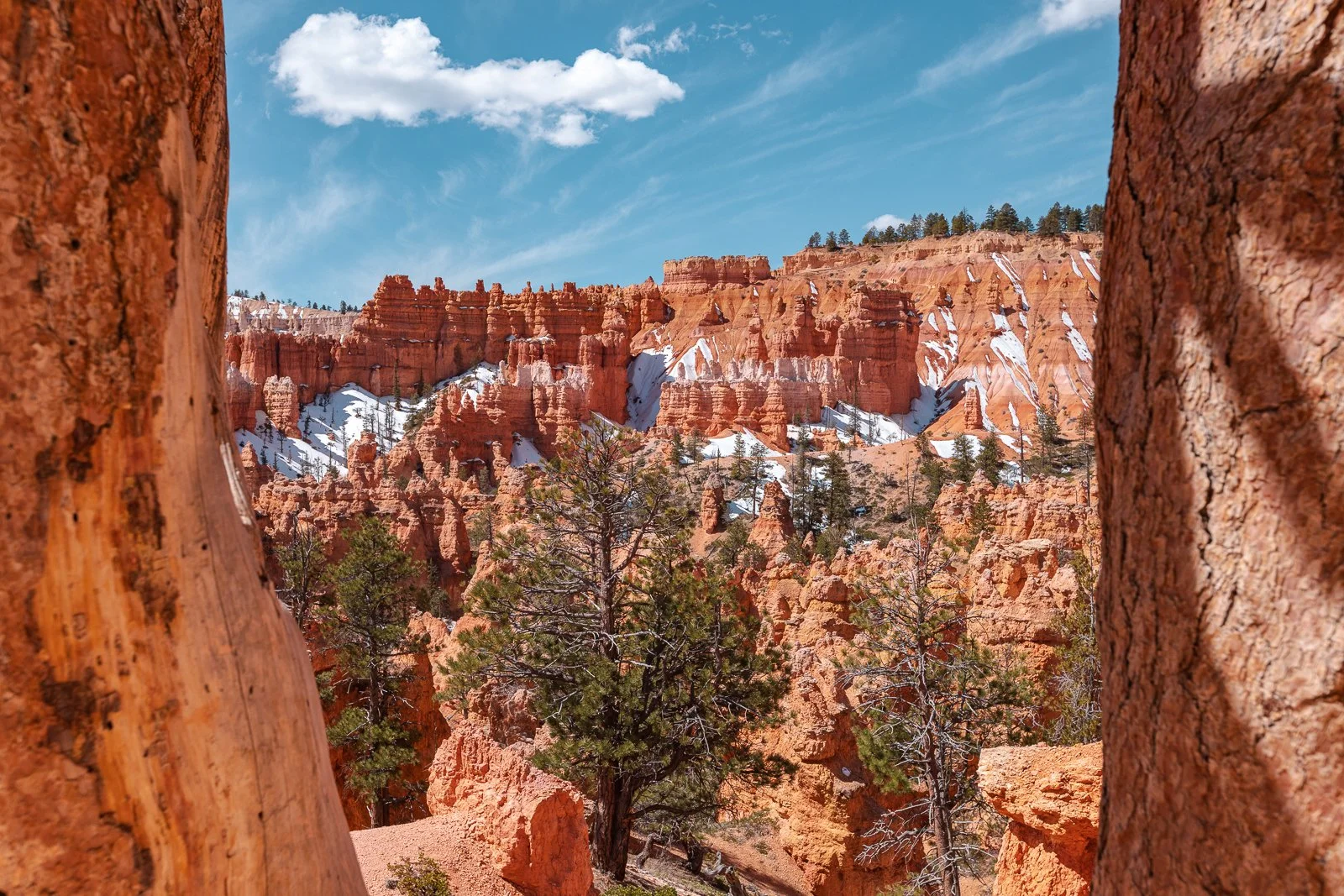 Paysage de Bryce Canyon avec formations rocheuses rouges, quelques arbres verts, ciel bleu avec quelques nuages, et traces de neige sur les rochers.
