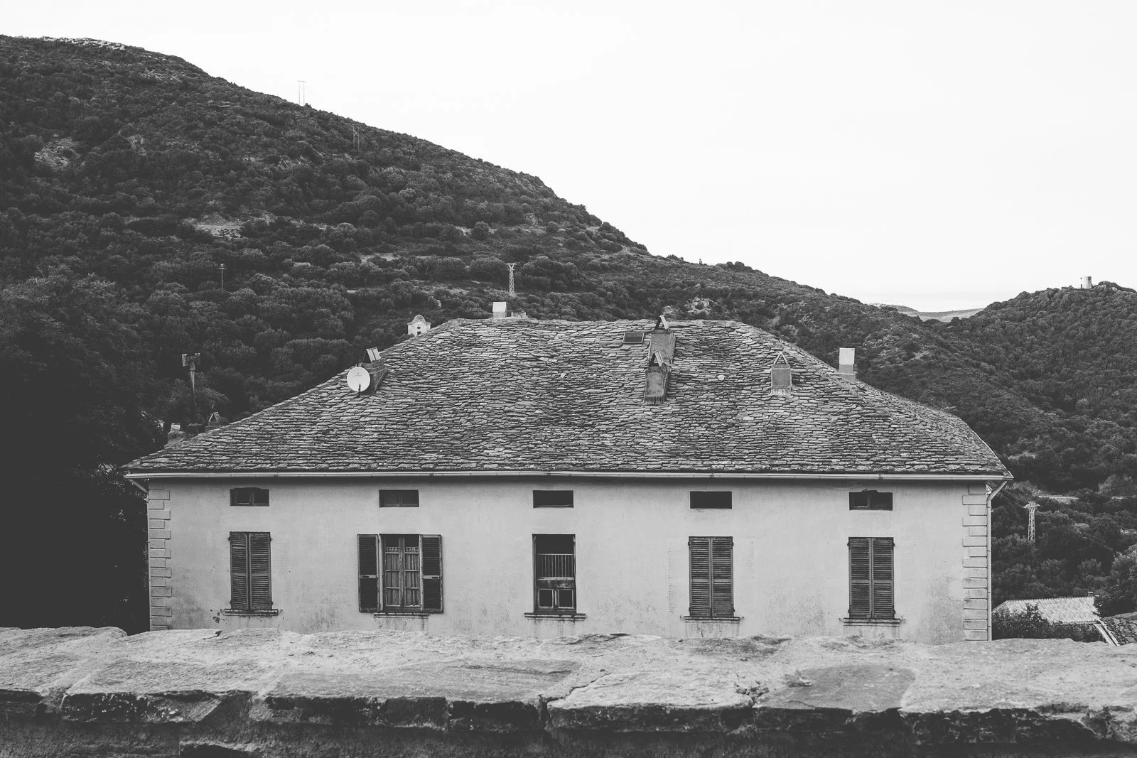 Une maison avec un toit en tuiles, fenêtres à volets et une cheminée, située dans un paysage de montagnes et de collines boisées.