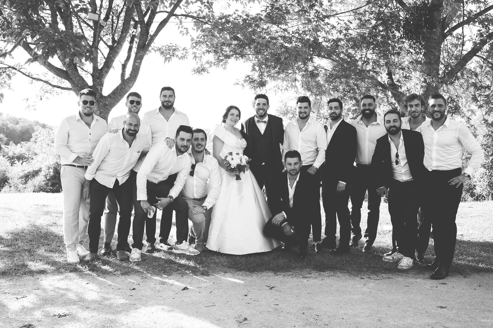 Groupe de personnes lors d'un mariage en plein air, comprenant un couple de jeunes mariés entourés de leurs amis, sous un arbre, en noir et blanc.
