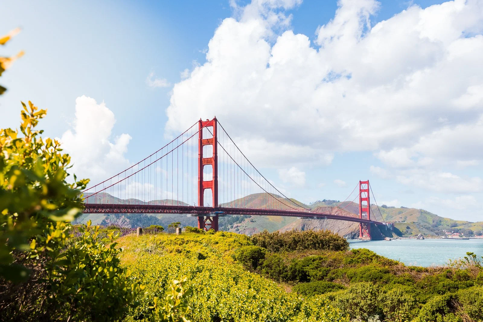 Vue du pont Golden Gate rouge sous un ciel partiellement nuageux, avec des collines verdoyantes et la côte au loin.