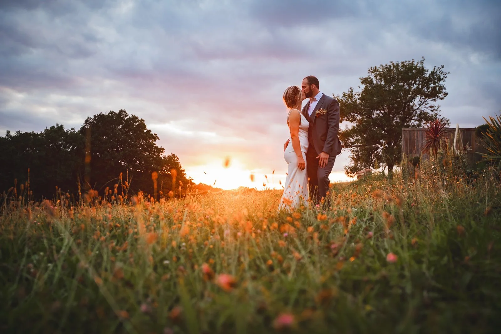 Un couple de mariés se tient dans un champ avec un coucher de soleil en arrière-plan, entouré de fleurs et d'arbres.