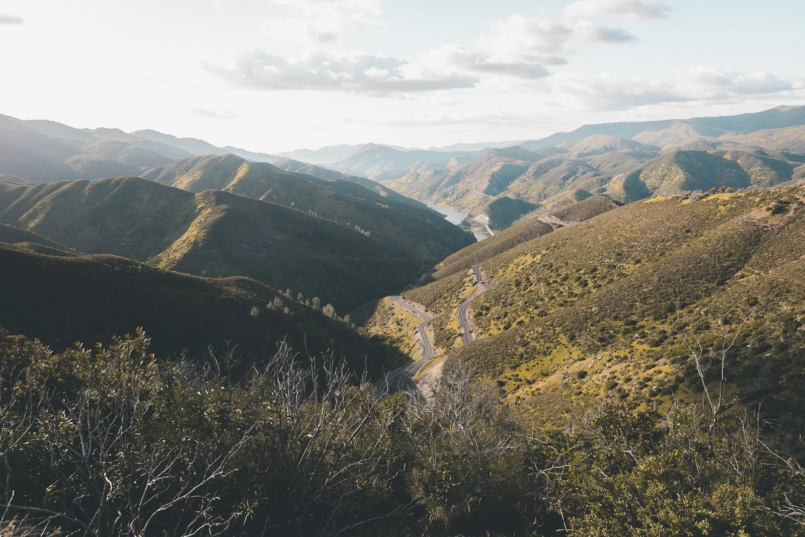 Paysage montagneux avec routes sinueuses, collines verdoyantes et un lac au fond, sous un ciel partiellement nuageux.