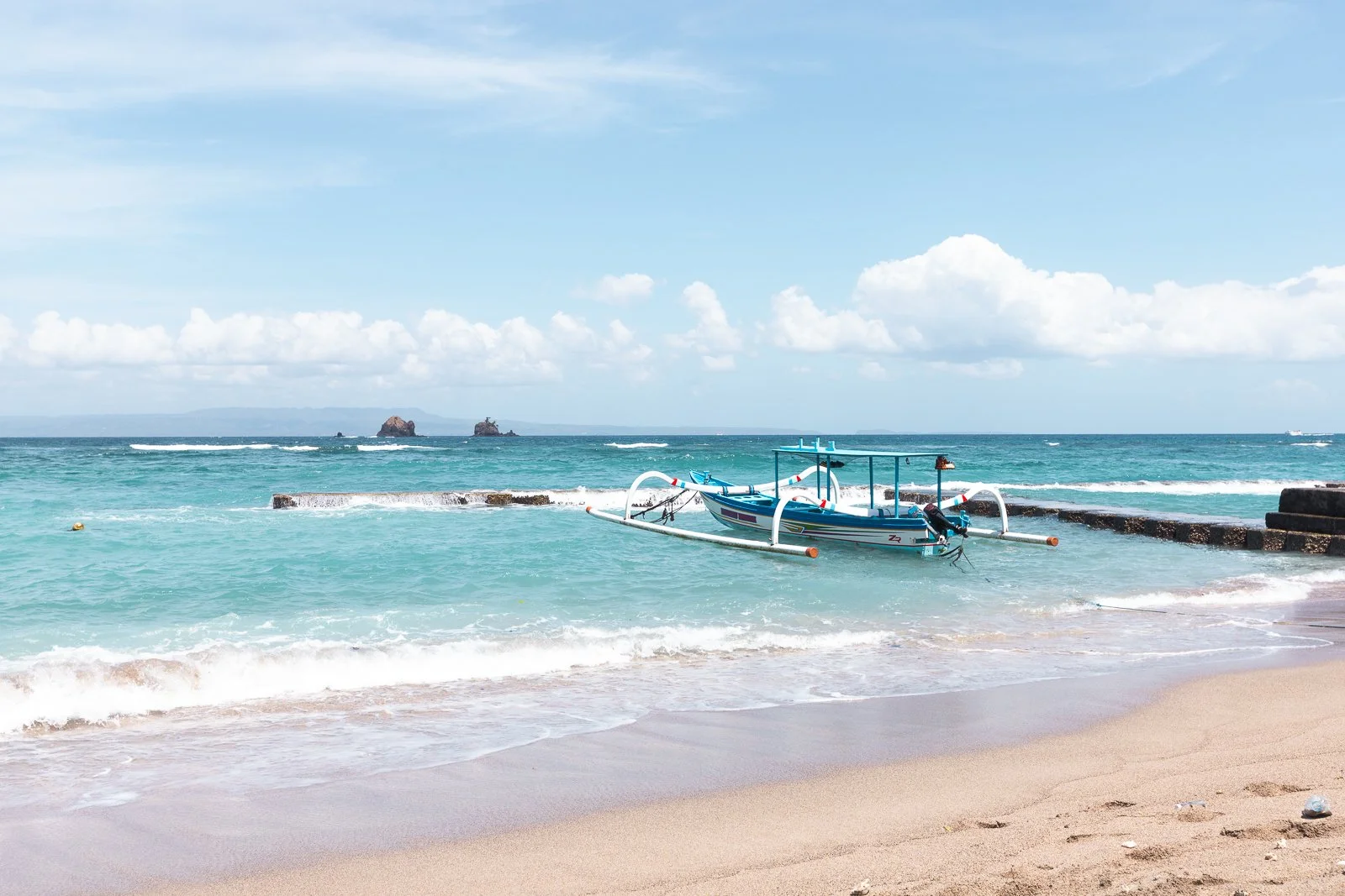 Plage de sable avec un bateau à rames coloré sur l'eau turquoise et des rochers au loin sous un ciel en partie nuageux.
