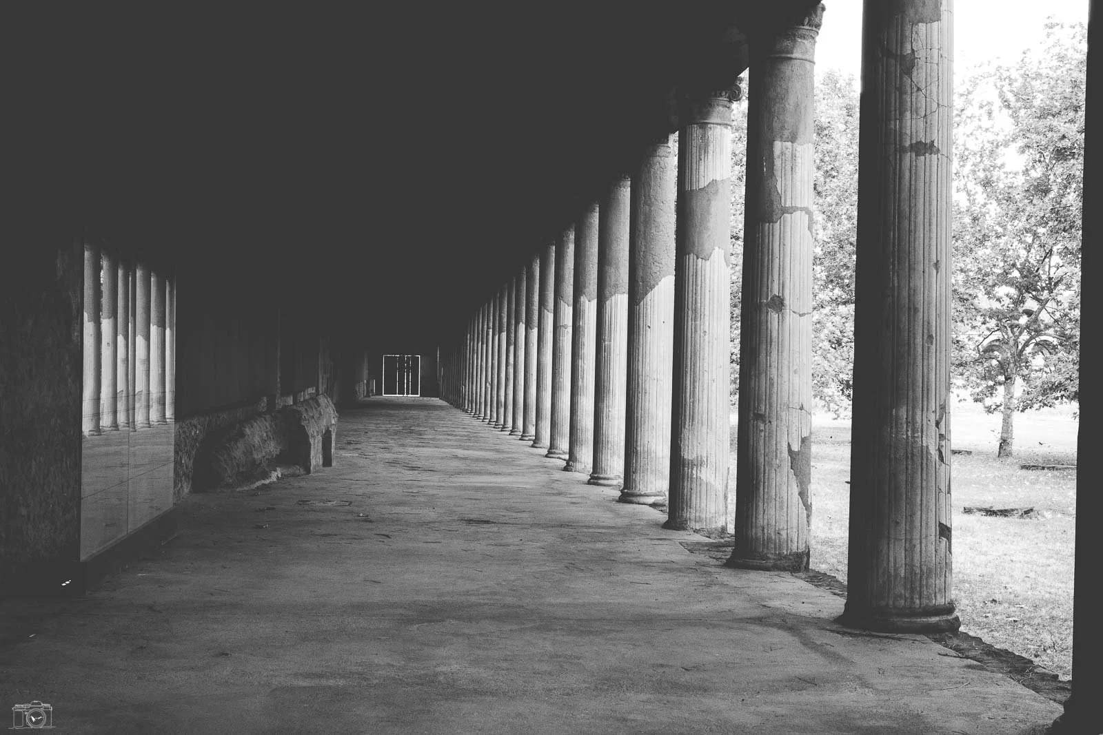 Un couloir avec des colonnes en pierre, dans un style antique, avec des arbres visibles à l'extérieur.