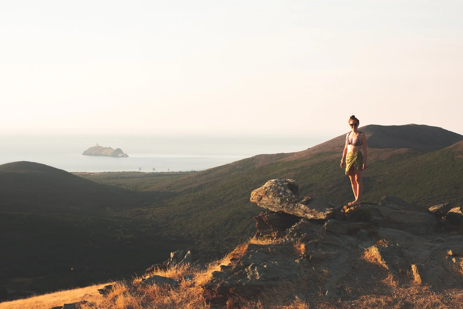 Une femme en maillot de bain et jupe en toile, portant des lunettes de soleil, se tient debout sur une roche en haut d'une colline avec une vue sur la mer, un îlot avec une statue ou un monument au loin.