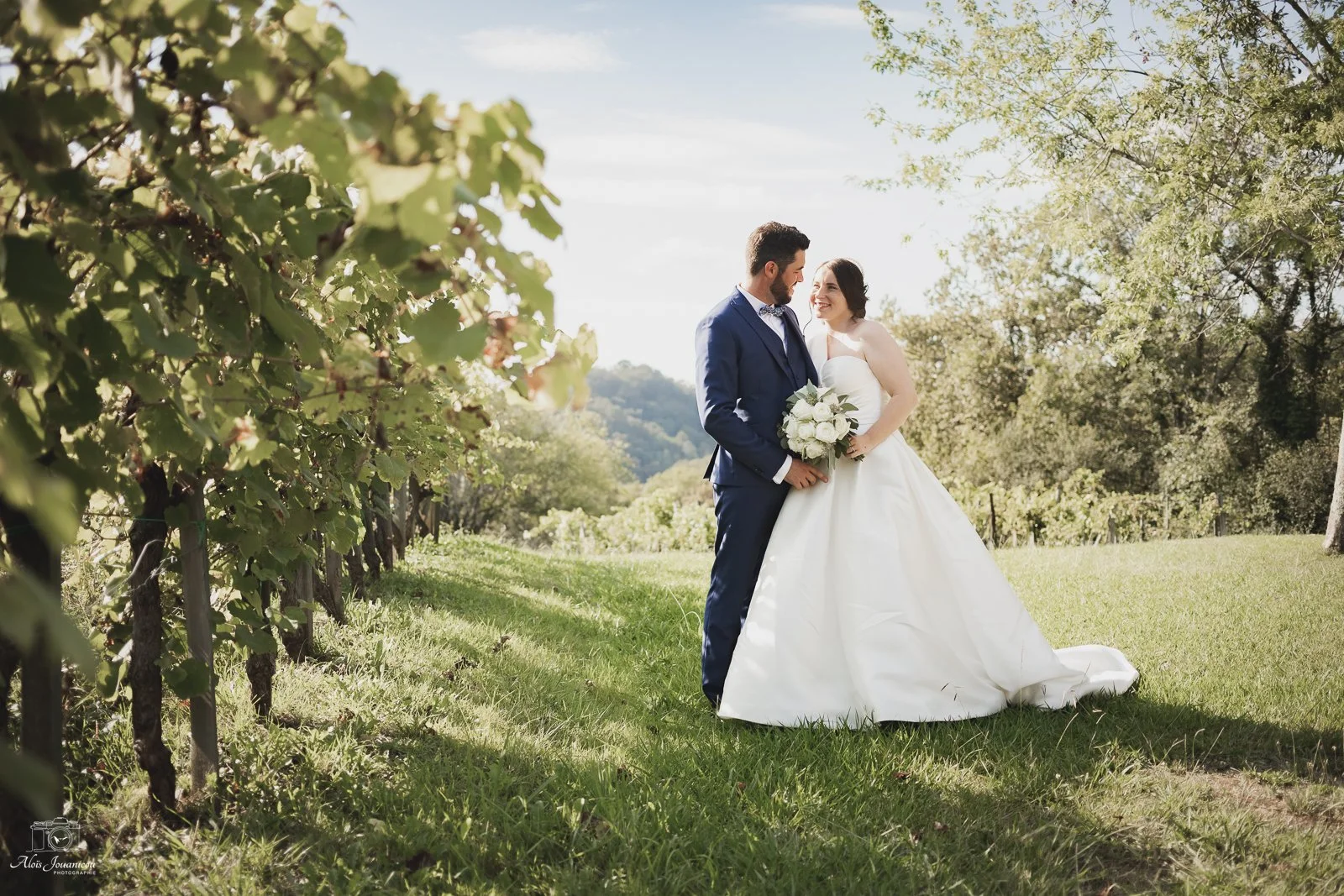 Un couple de mariés se tenant la main dans un vignoble en plein air lors de leur mariage, avec des arbres et des collines en arrière-plan.