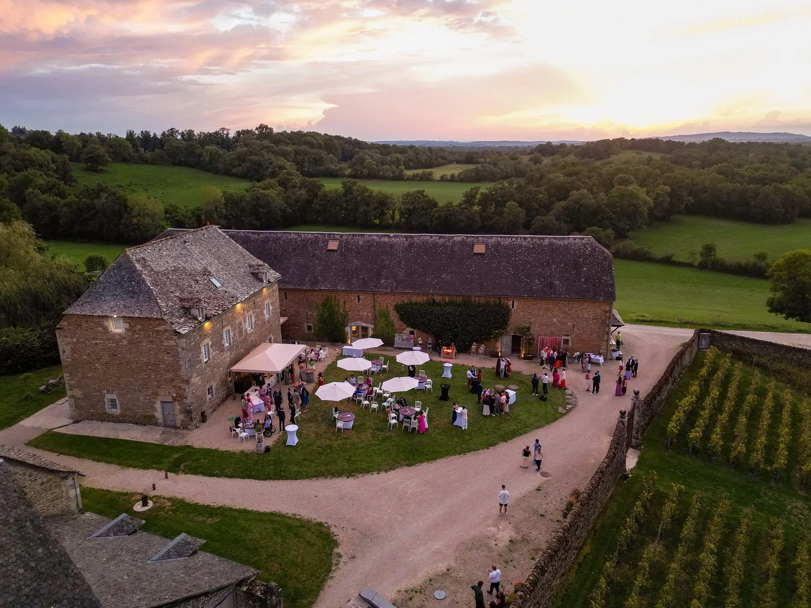Soirée de mariage avec des invités dans une ferme en campagne, plusieurs tables avec parasols, un grand bâtiment en pierre, un ciel coloré au coucher du soleil, forêt et champs verts environnants.