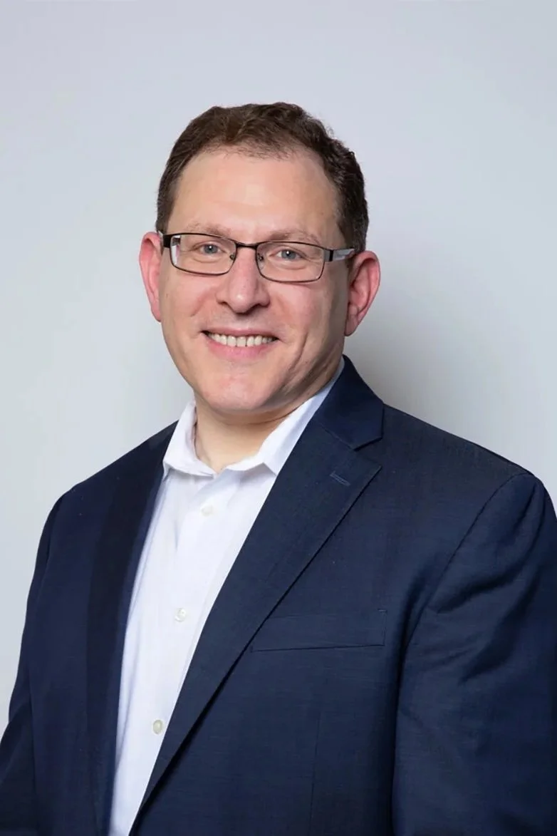 A man in a suit smiling at the camera, wearing glasses, against a plain white background.