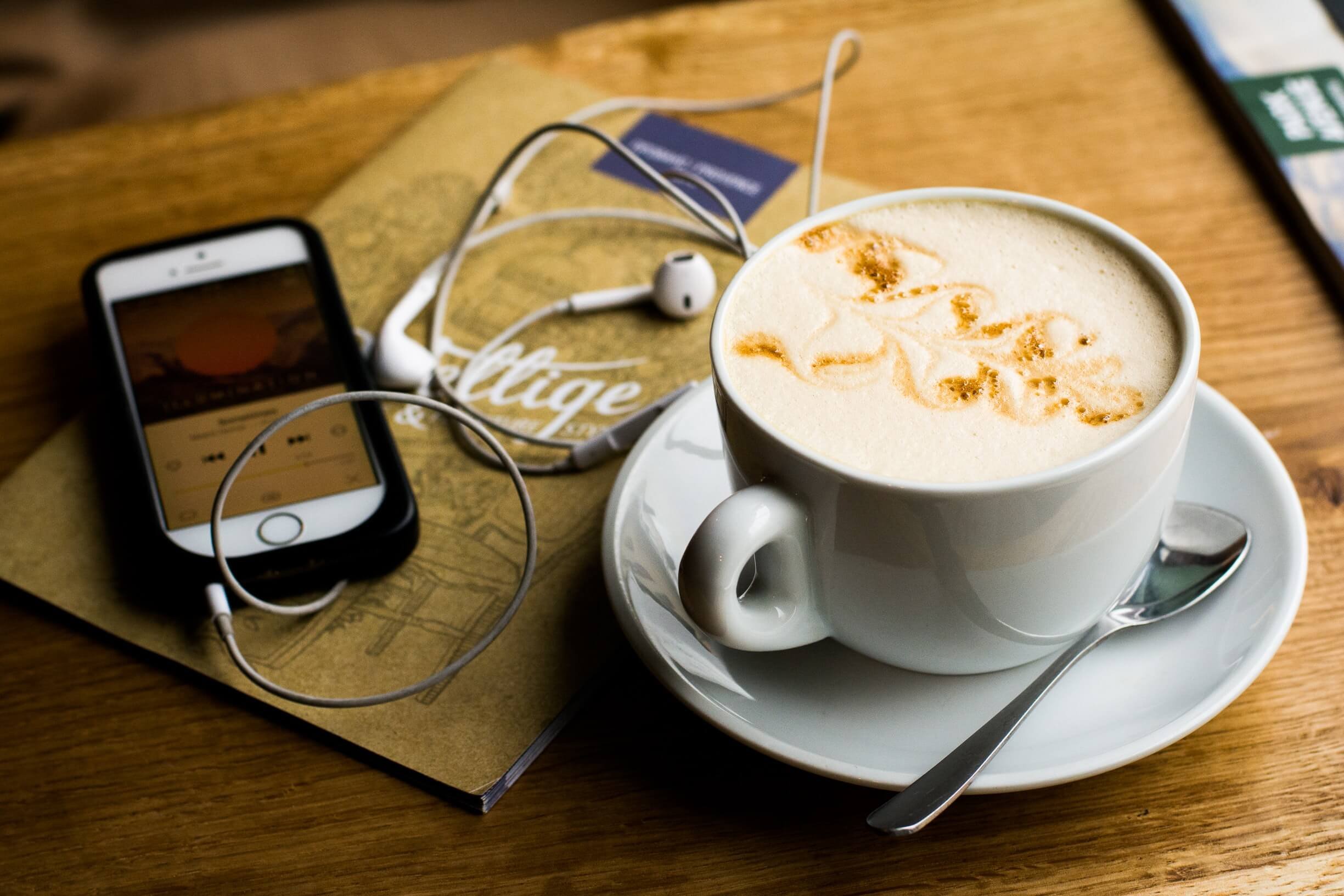 A cup of coffee with foam and caramel syrup, a spoon, a smartphone, white earphones, a magazine, and a silver pen on a wooden table.