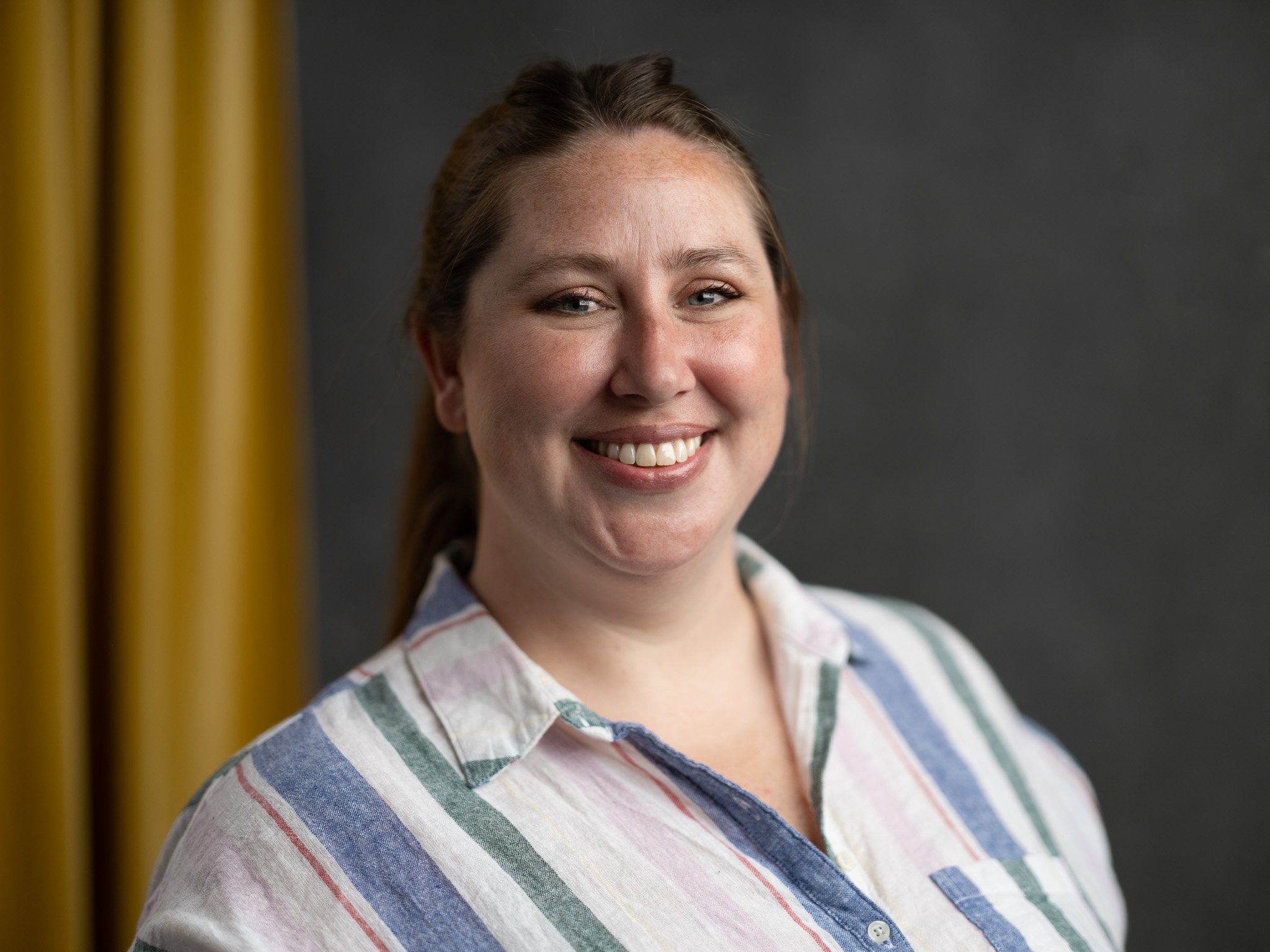 Close-up of a young woman smiling, wearing a colorful striped shirt, in front of a dark background with yellow curtains.