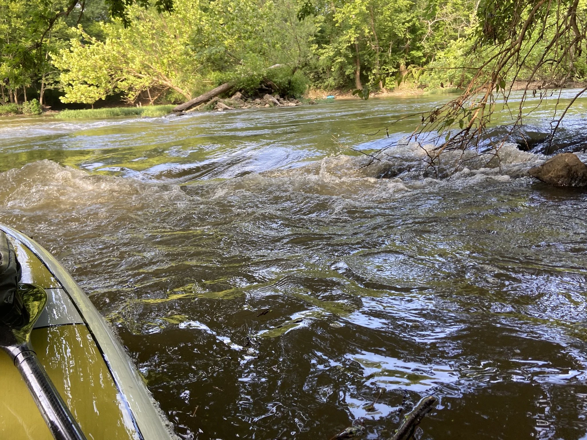 View of a river with flowing water and surrounding green trees, seen from a kayak or small boat in the foreground on the left side.