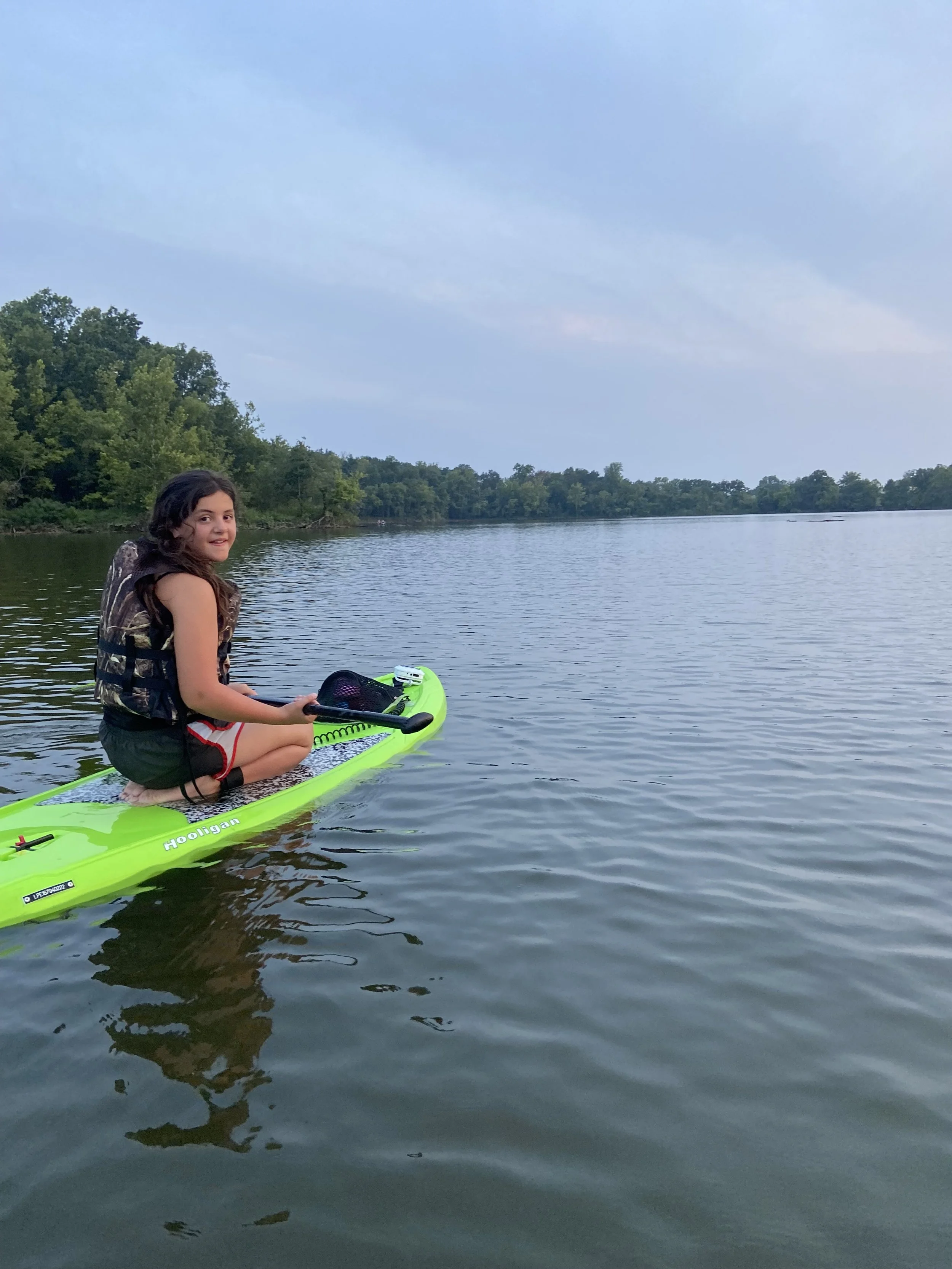 A young girl sitting on a bright green paddleboard on a calm lake, wearing a life jacket. She is holding a paddle and smiling, with trees lining the distant shore under an overcast sky.