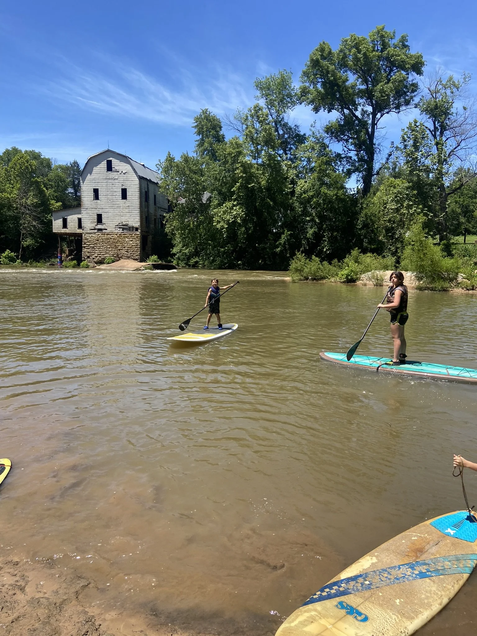 People paddleboarding on a river with a rustic gris mill and green trees in the background under a blue sky.